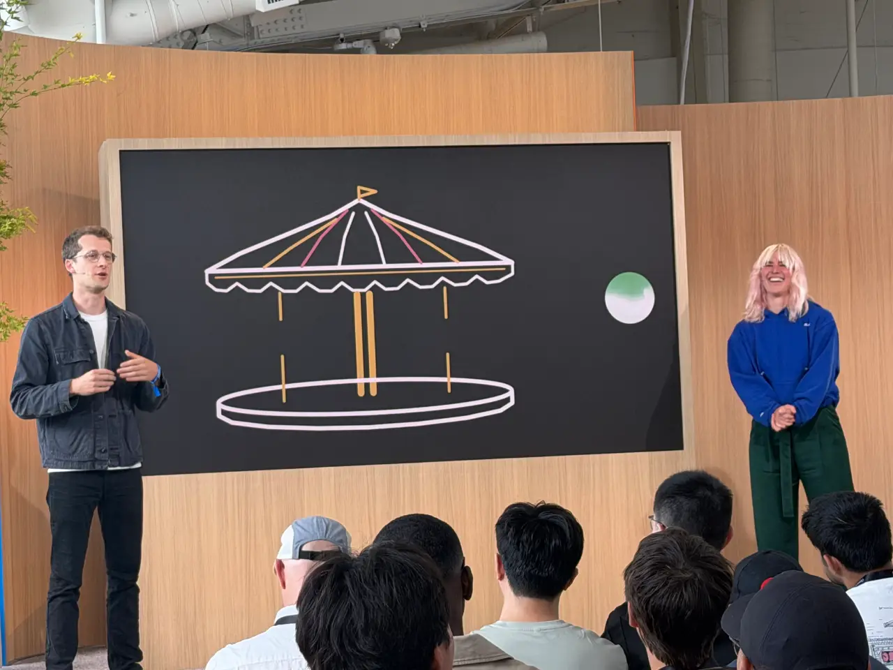 Two presenters address a seated audience in a wood-paneled venue, standing on either side of a large screen showing a colorful line drawing of a carousel and a green-and-white sphere.